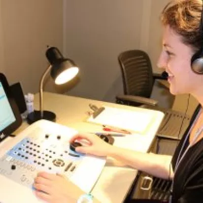Graduate student on right with arms extended to another person's ear, fitting for hearing equipment