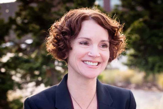 Smiling woman with short, curly auburn hair wears a dark blazer over a burgundy top.  She has a tree-of-life pendant necklace.  Outdoor setting.